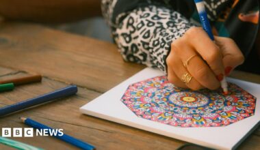 A person draws a colourful round pattern using pencils and pens. They are wearing a white and black animal print shirt and gold rings.