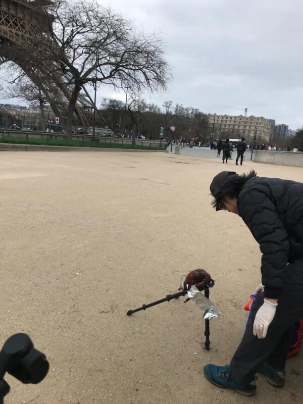 A person wearing gloves bends over next to a tripod holding a bird puppet or sculpture near the Eiffel Tower in Paris. Several people walk in the background on an overcast day.