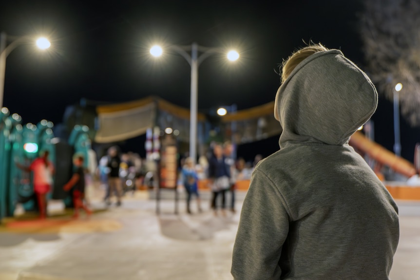 A young boy sits with his back towards us wearing a hooded jumper, looking up at the night sky with lights and people around.