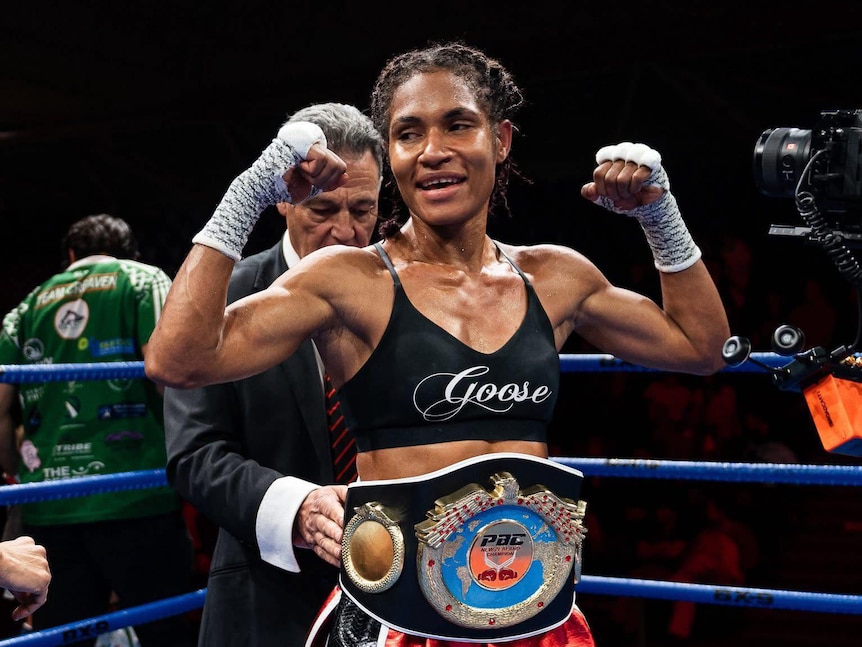 A young Papua New Guinean has a championship boxing belt put around her waist.  She smiles and has her hands up in the air