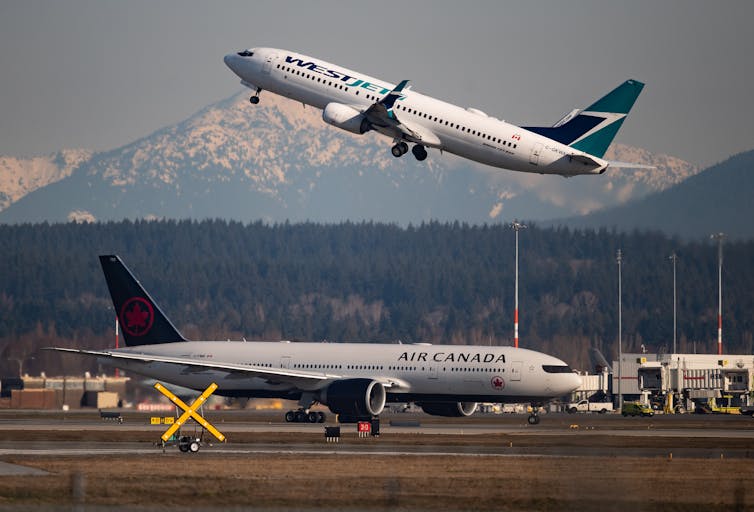 An airport tarmac with a plane taxiing in the foreground and one taking off in the background
