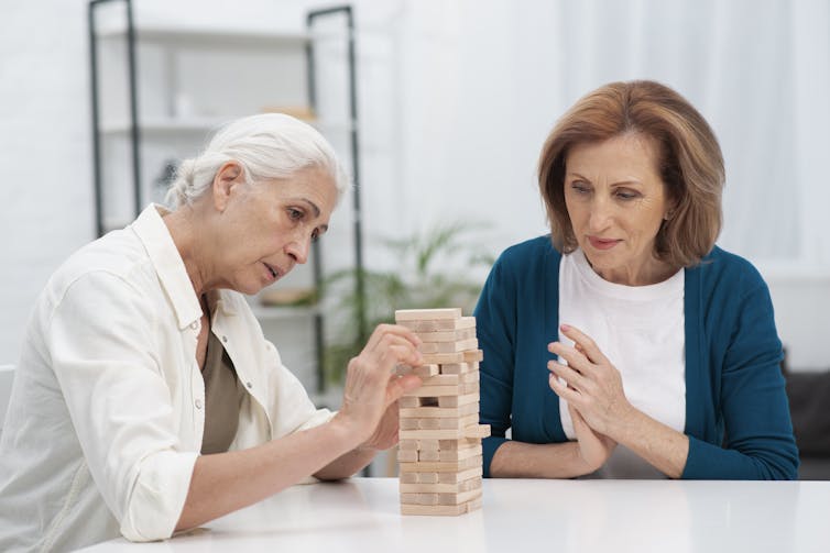 A woman with grey hair doing a puzzle while a younger woman observes.