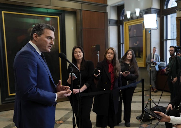 A man in a blue suit standing at a microphone with other people in the background