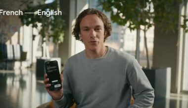 A man in a gray sweater holds up a phone that's doing live translation of French to English language. He's wearing earbuds.