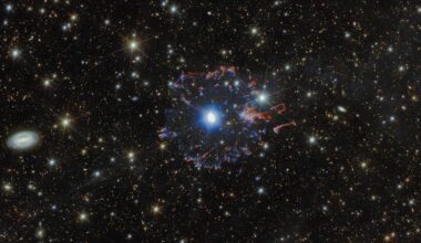 A planetary nebula in space. The star in the very center is surrounded by white bubbles and loops of gas, all shining with a powerful blue light. Farther away a broken ring of red and blue gas clouds surrounds the nebula. A multitude of golden and white stars, wisps of gas and distant galaxies of various sizes surround the nebula on the black background.
