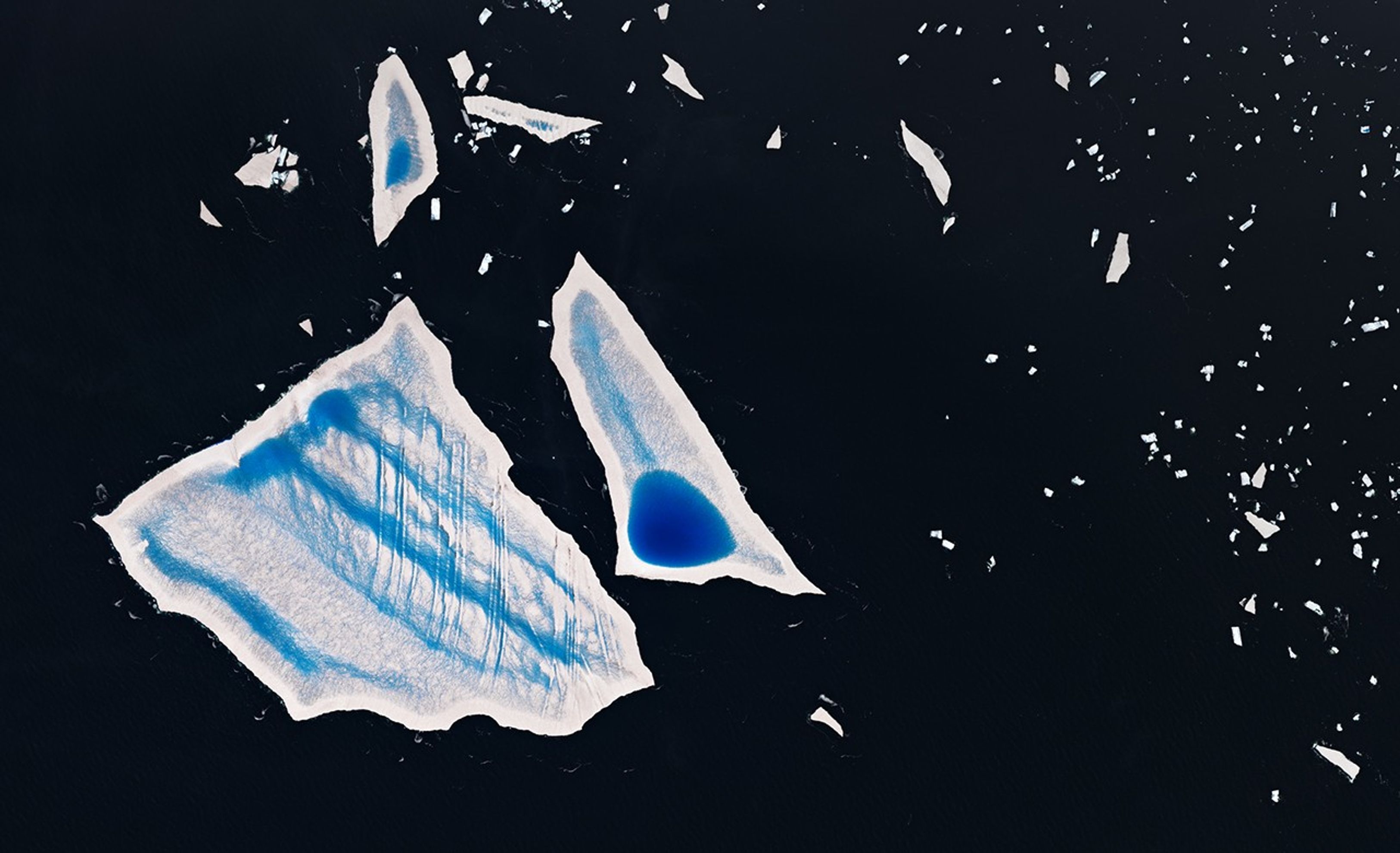 A more detailed view of large fragments of A-23A shows distinct melt pools and channels on the surfaces of irregularly shaped icebergs against dark ocean waters. Dozens of much smaller icebergs are scattered around the largest bergs, particularly on the right side of the image.