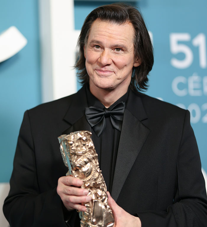 Jim Carrey holding an award, dressed in a black suit and bow tie at a formal event with a blue background. Jim Carrey holding an award, dressed in a black suit and bow tie at a formal event with a blue background.