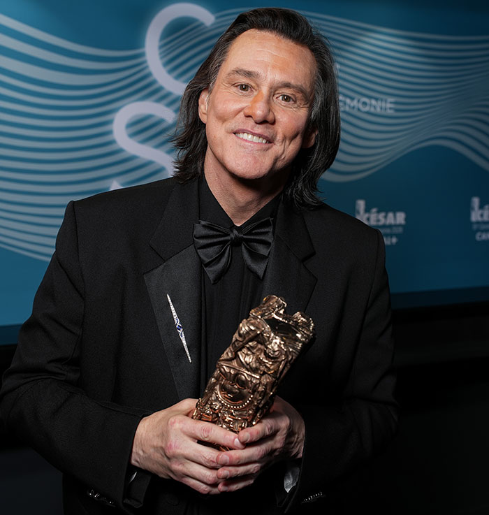 Man resembling Jim Carrey in black tuxedo holding an ornate trophy at an award event with wavy blue backdrop. Man resembling Jim Carrey in black tuxedo holding an ornate trophy at an award event with wavy blue backdrop.