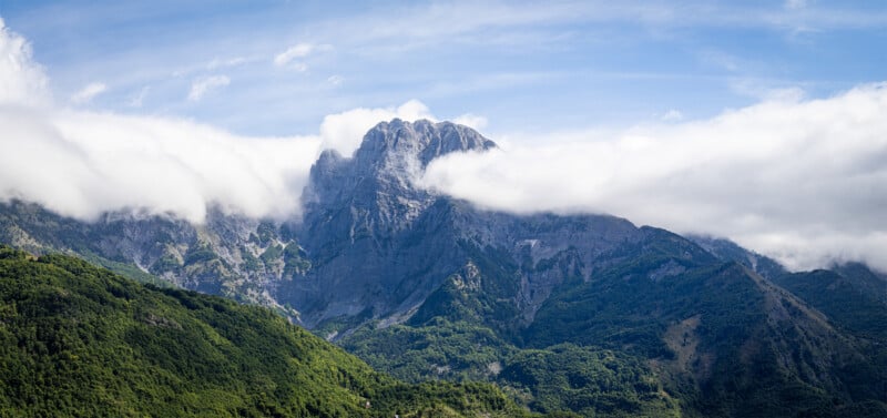 A towering rocky mountain peak partly covered by clouds rises above lush green hills under a blue sky with wispy clouds.