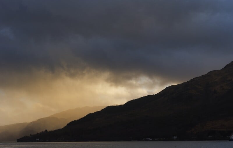 A dramatic sky with dark clouds over a mountain range at sunset, golden light breaking through and illuminating the landscape, with a calm body of water in the foreground.