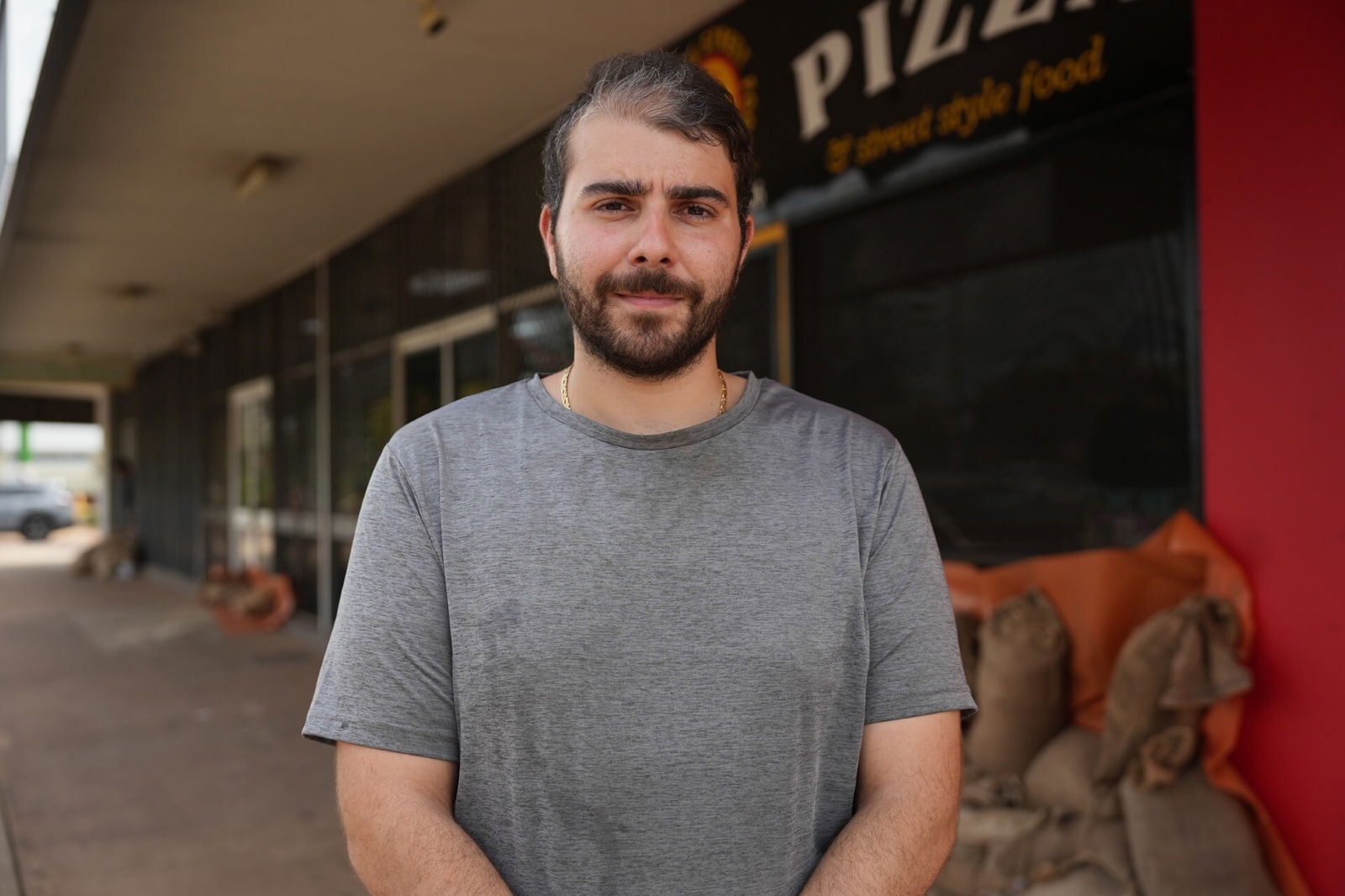 A white man, black hair with grey streak, brown hear, wearing gray shirt, standing in front of his pizza shop.