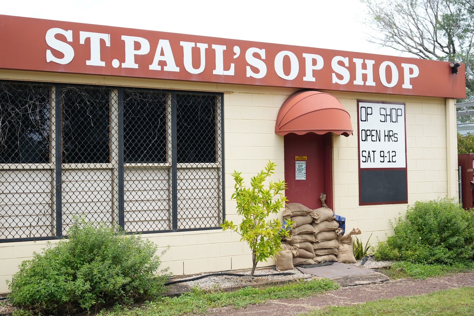 A St Paul's Op Shop, orange sign, white building, sandbags piled in front of it's front red door.