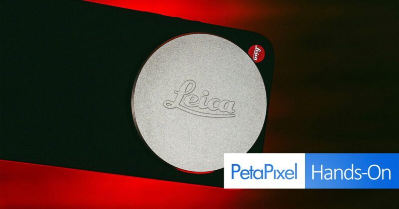 Close-up of a Leica camera lens cap with the Leica logo embossed on it, set against a dark background with a red glow. A "PetaPixel Hands-On" label appears at the bottom right corner.