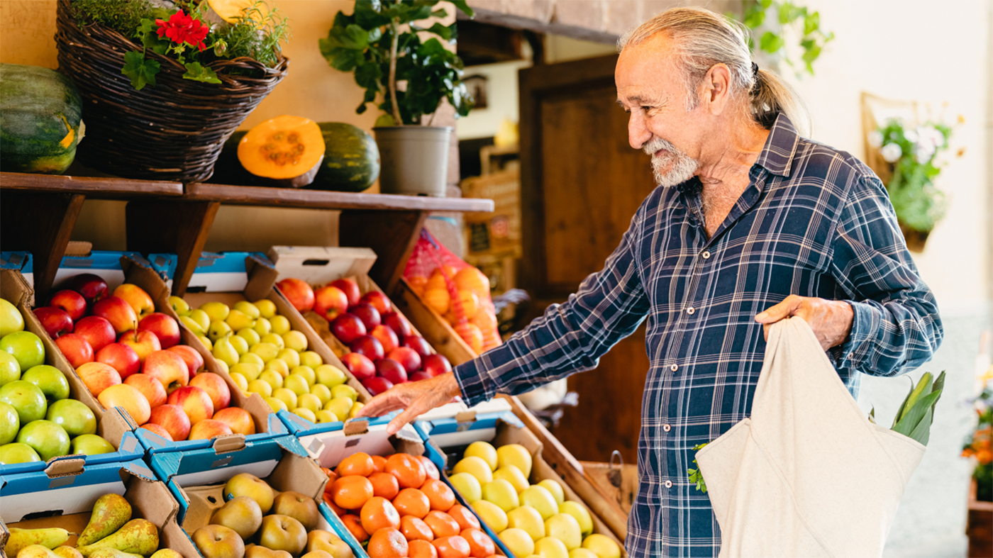 Elderly man in a plaid shirt smiling while selecting fruits from a market stand during National Nutrition Month. Vibrant apples, pears, and tomatoes are displayed in boxes nearby.