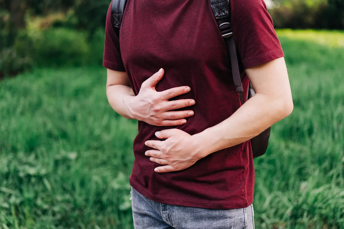 A person wearing a red shirt holds their stomach, standing outdoors in a grassy area.