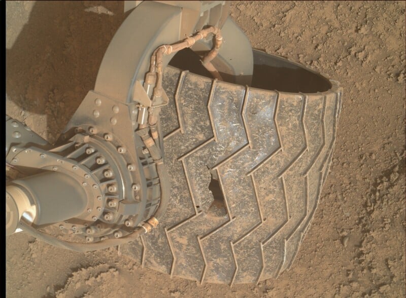 Close-up of a rover’s metal wheel with visible cracks and dents on its surface, resting on dusty, reddish Martian soil. The mechanical joint and wiring are also visible.