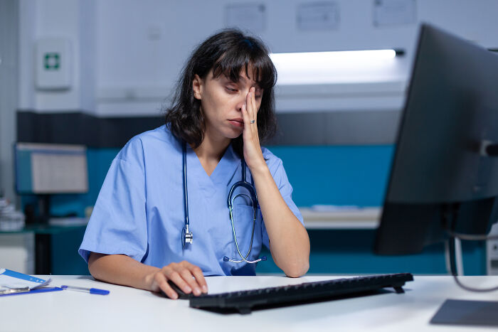 Female doctor in scrubs with stethoscope, looking stressed and tired while working late on a computer in a medical office.