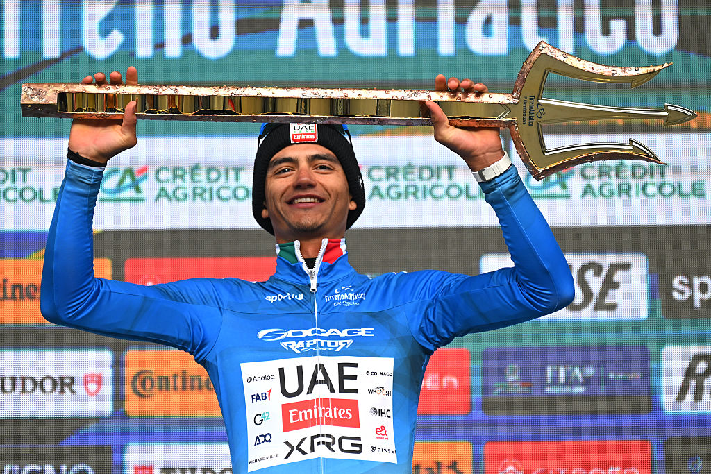 Isaac del Toro on the podium wearing the blue jersey and holding the trident trophy of the race winner overhead