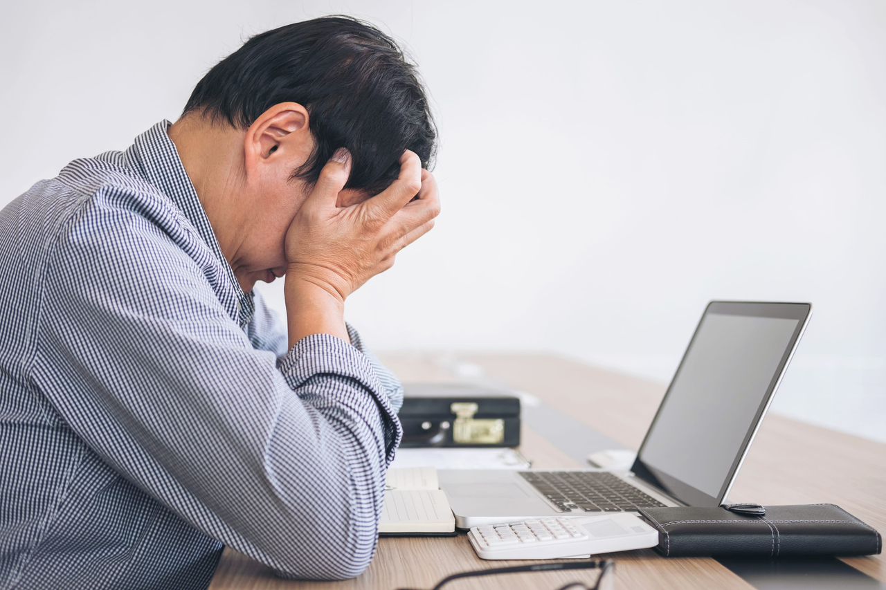 A man holds his head while sitting at a desk with a laptop. (123rf)