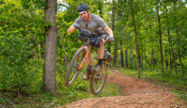A mountain biker performing a jump on a dirt trail surrounded by trees, smiling as he propels off the ground with his bike. The biker is wearing a helmet, gloves, and casual clothing, and the terrain is a mix of gravel and natural foliage.