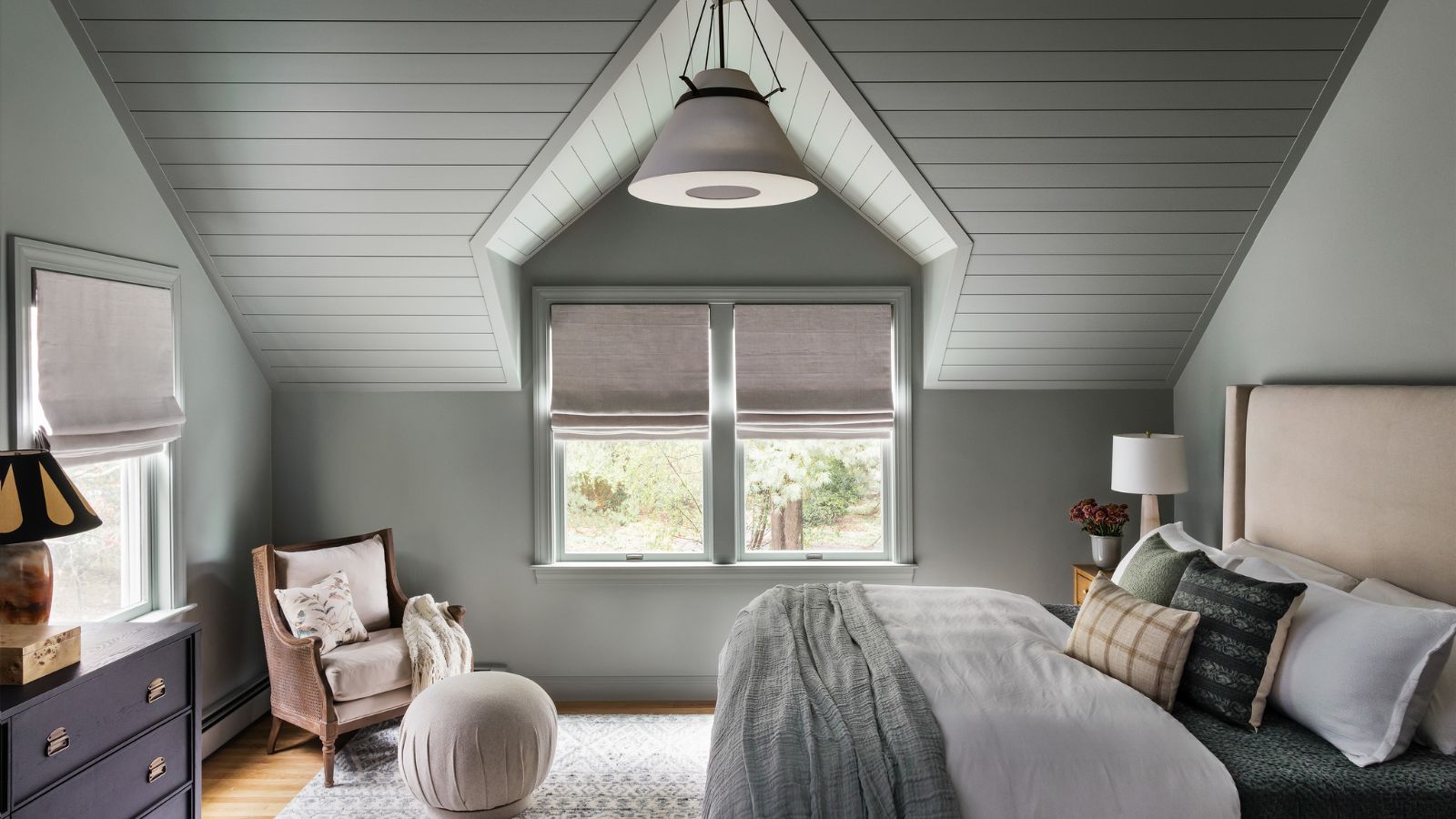A bedroom with color-drenched pale green walls and a sloped ceiling with panelling. A large taupe headboard and layered neutral-colored bedding, cushions, and throws.
