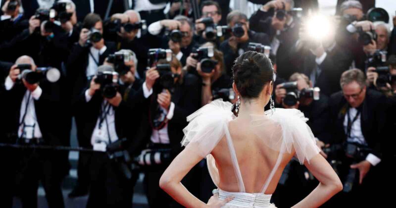 A woman in a glamorous dress poses with her back to the camera on a red carpet, facing a crowd of photographers taking pictures with bright flashes.
