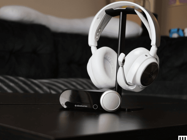 White headset with cushioned ear cups and extended mic hooked on a tabletop stand. Oval-shaped device to customize the audio is in front of the stand.