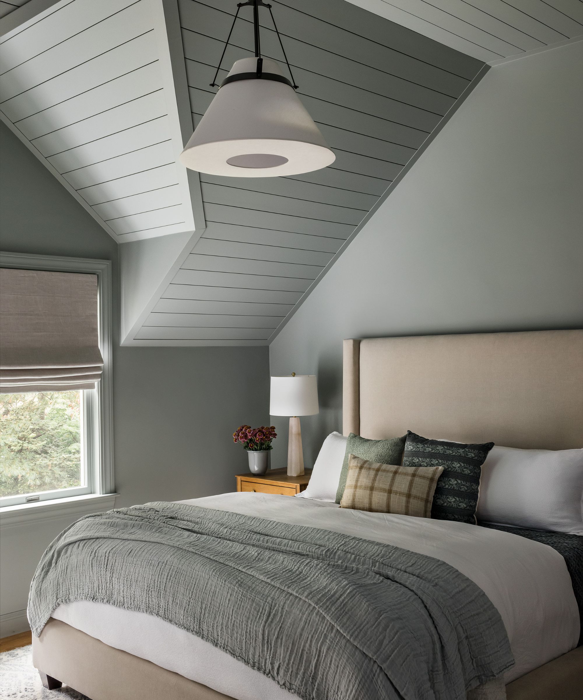 A bedroom with color-drenched pale green walls and a sloped ceiling with panelling. A large taupe headboard and layered neutral-colored bedding, cushions, and throws.