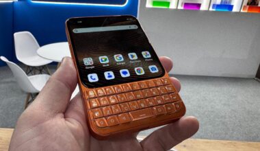 A hand holding a small-format orange phone with a full but compact QWERTY keyboard under a square phone screen with a modern Android interface.