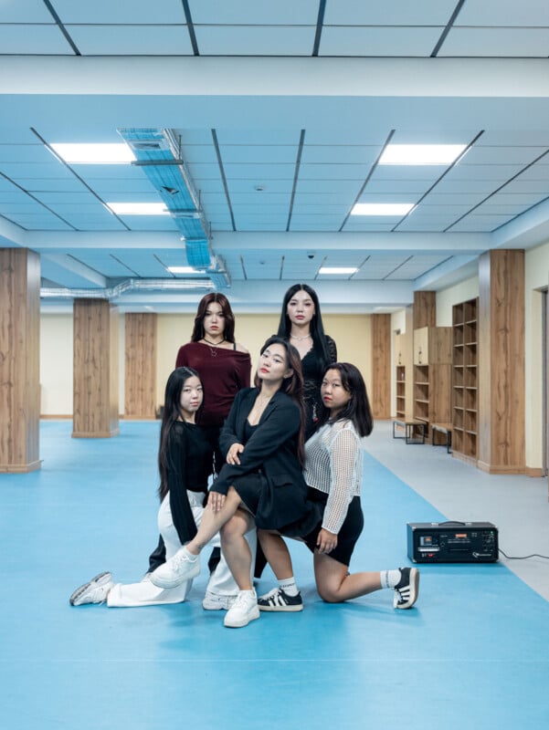 Five young women pose confidently in a spacious room with blue floors, wood pillars, and a tiled ceiling. Some are kneeling, others standing, all facing the camera. A black speaker sits on the floor to the right.
