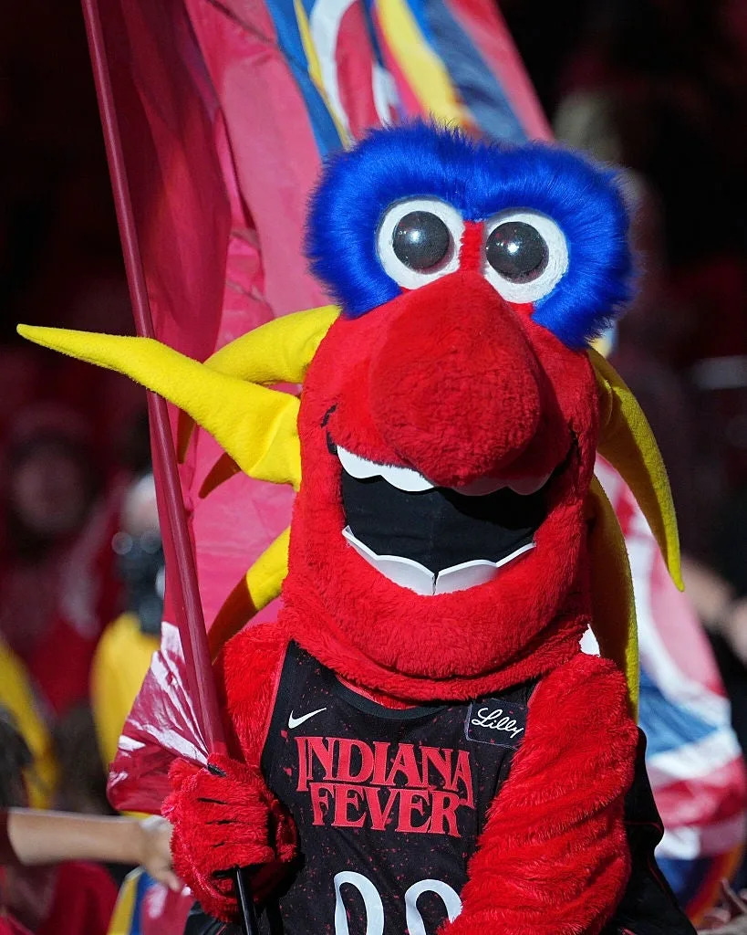 A red and yellow mascot with a large feathered head holds a flag at a sports event, wearing an Indiana Fever jersey