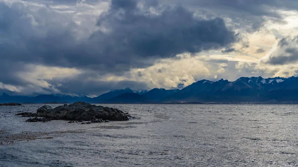 A tiny rocky islet in the ocean. Ripples on the water, algae on the surface. In the distance, the Andes mountain range is visible against the sky and dark clouds. Argentina. Tierra del Fuego