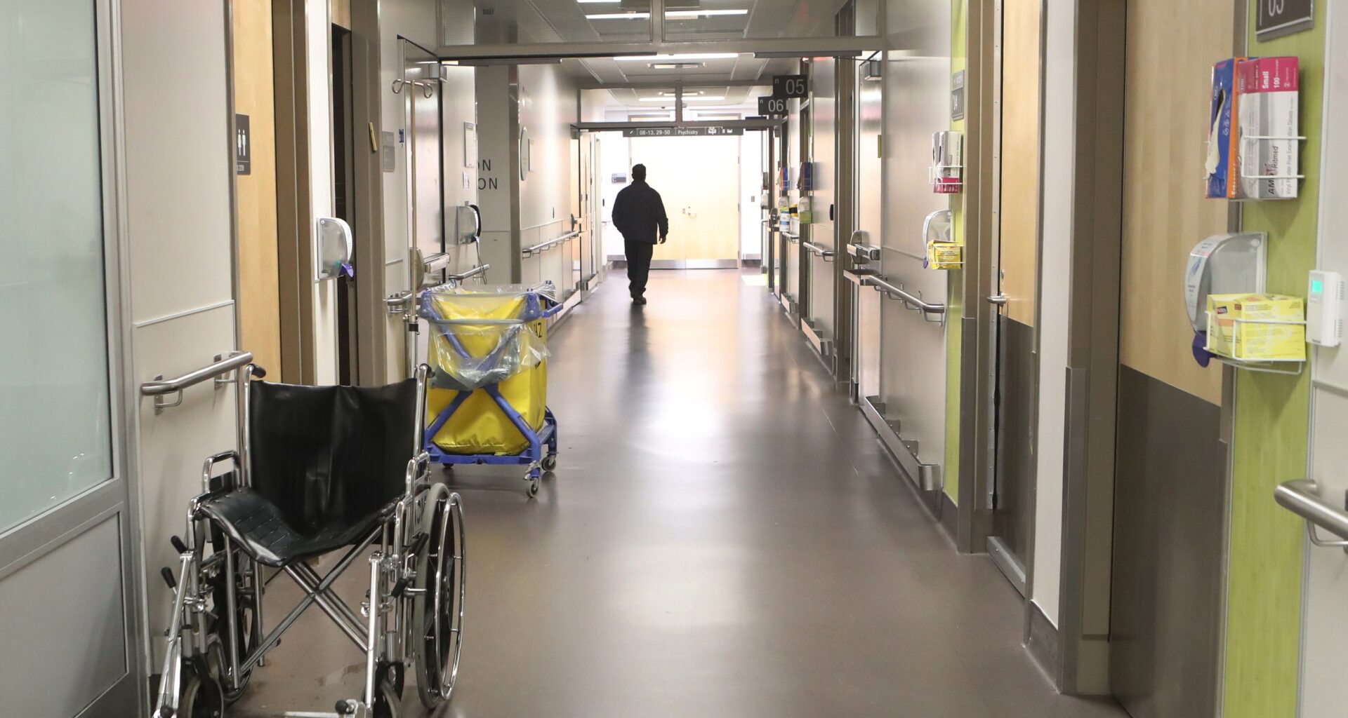 A hospital hallway with light coming through a window at the end. There is a wheelchair to the left in the foreground and a person in the distance, walking away.