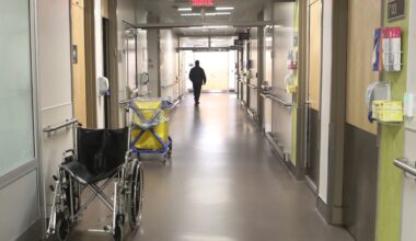 A hospital hallway with light coming through a window at the end. There is a wheelchair to the left in the foreground and a person in the distance, walking away.