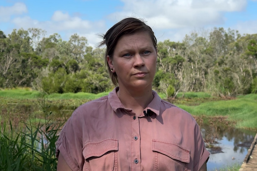 A woman with short brown hair, wearing a pink shirt stands in front of wetlands.
