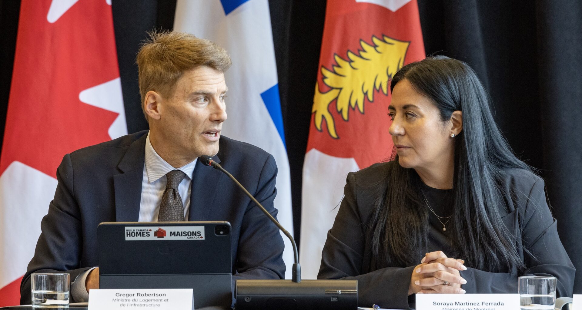 Soraya Martinez Ferrada and Gregor Robertson look at each other while sitting at a table with Canadian, Quebec and Montreal flags behind them.