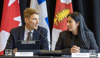 Soraya Martinez Ferrada and Gregor Robertson look at each other while sitting at a table with Canadian, Quebec and Montreal flags behind them.