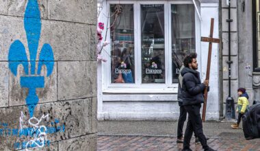 A man carries a large crucifix down a cobblestone street past a wall with a blue fleur-de-lys painted on it.
