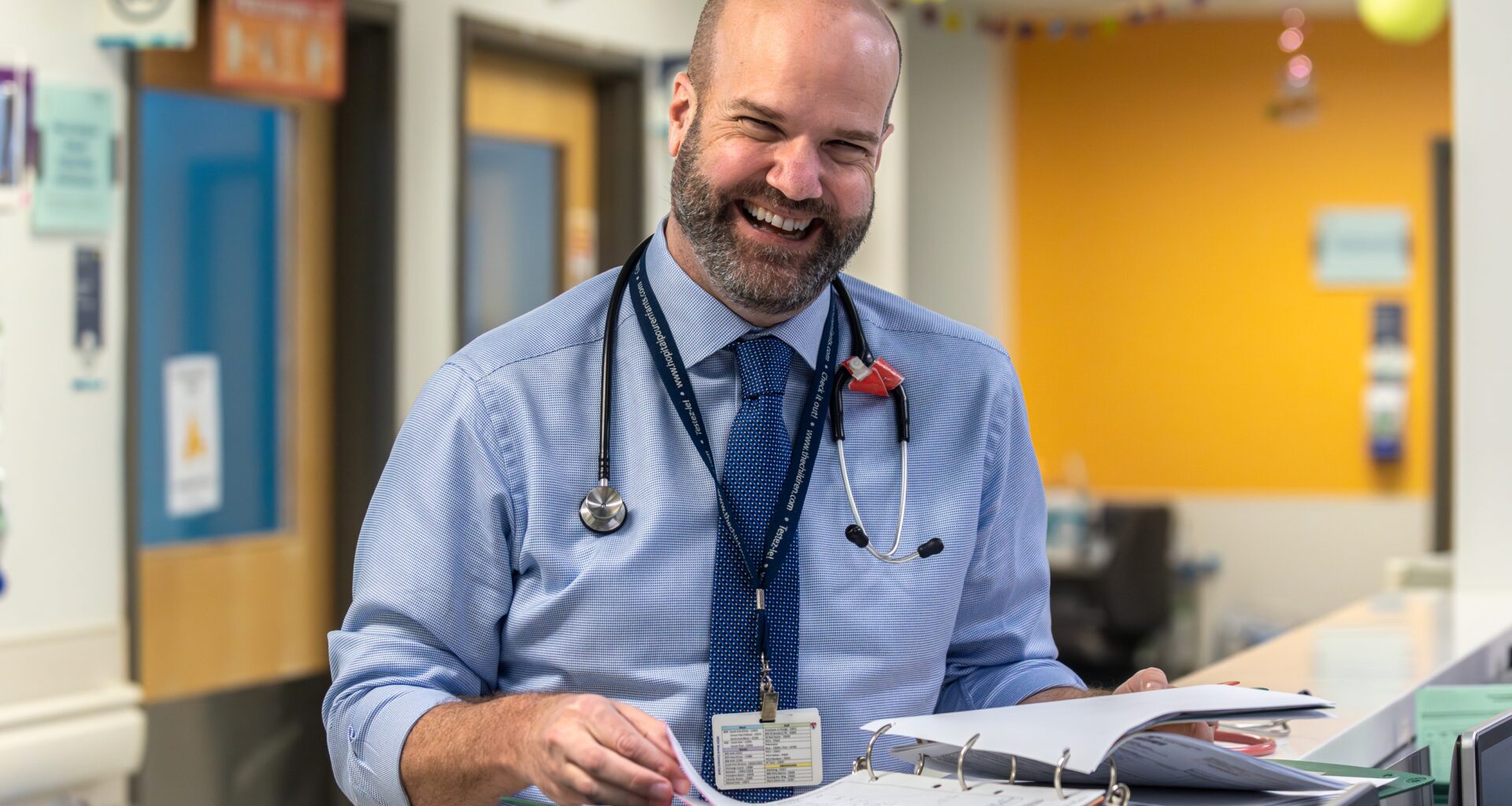 A doctor laughs while looking up from a medical file in the hallway of a hospital.