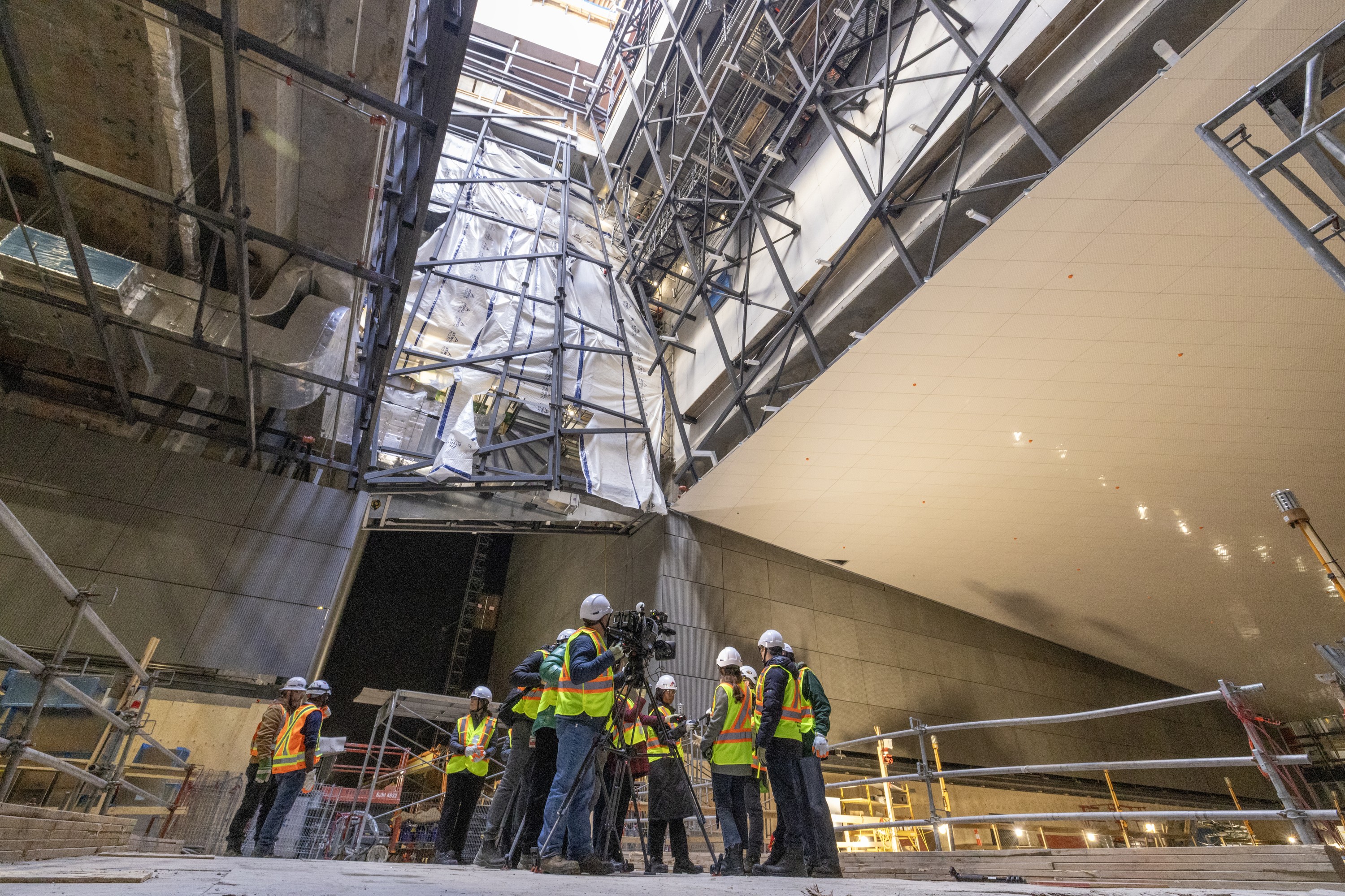 People in construction gear and with media cameras stand on a platform in a construction site, with light coming from above.