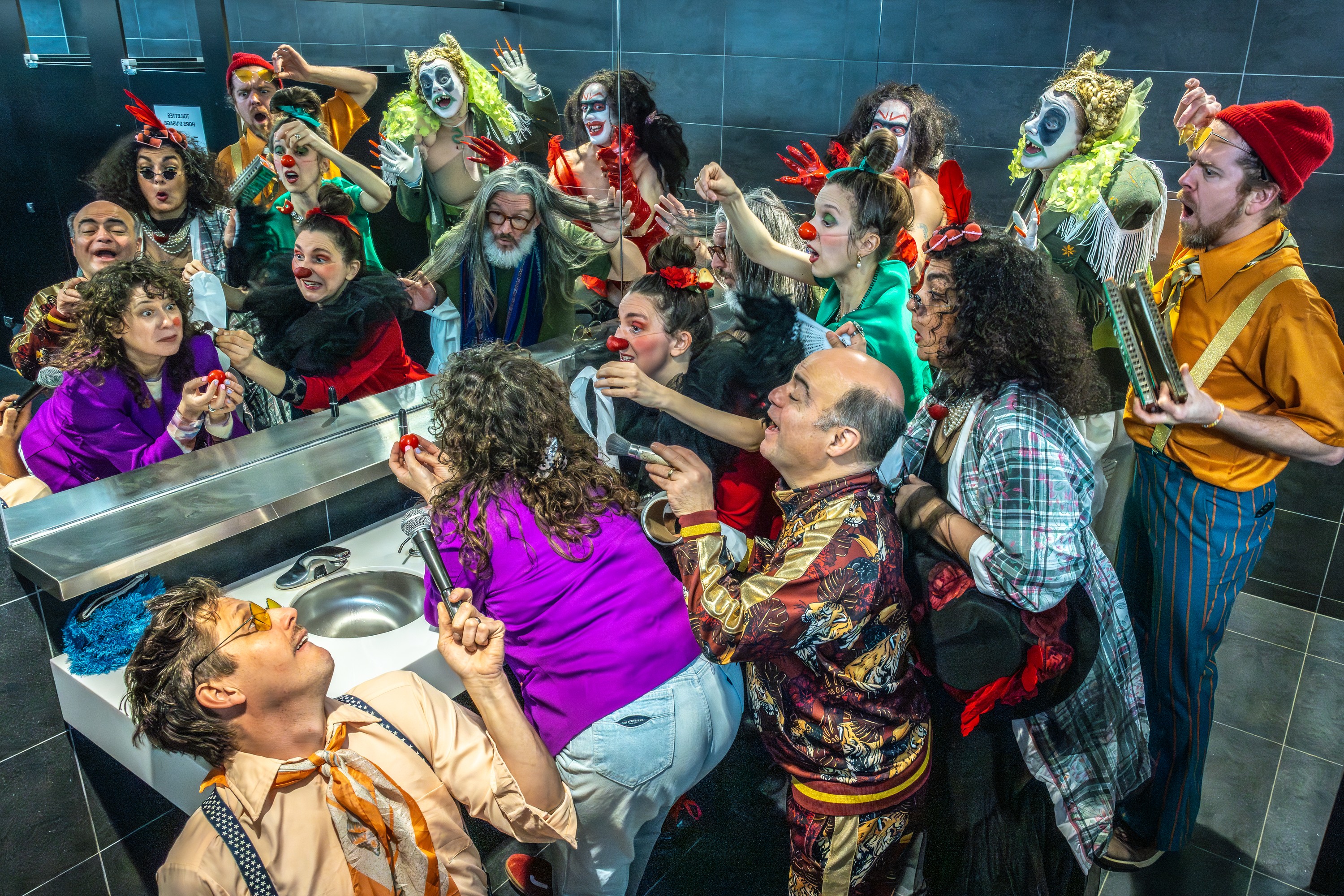 A woman is about to put on a clown nose as she looks in the mirror in a bathroom, surrounded by other people in clown outfits and makeup.