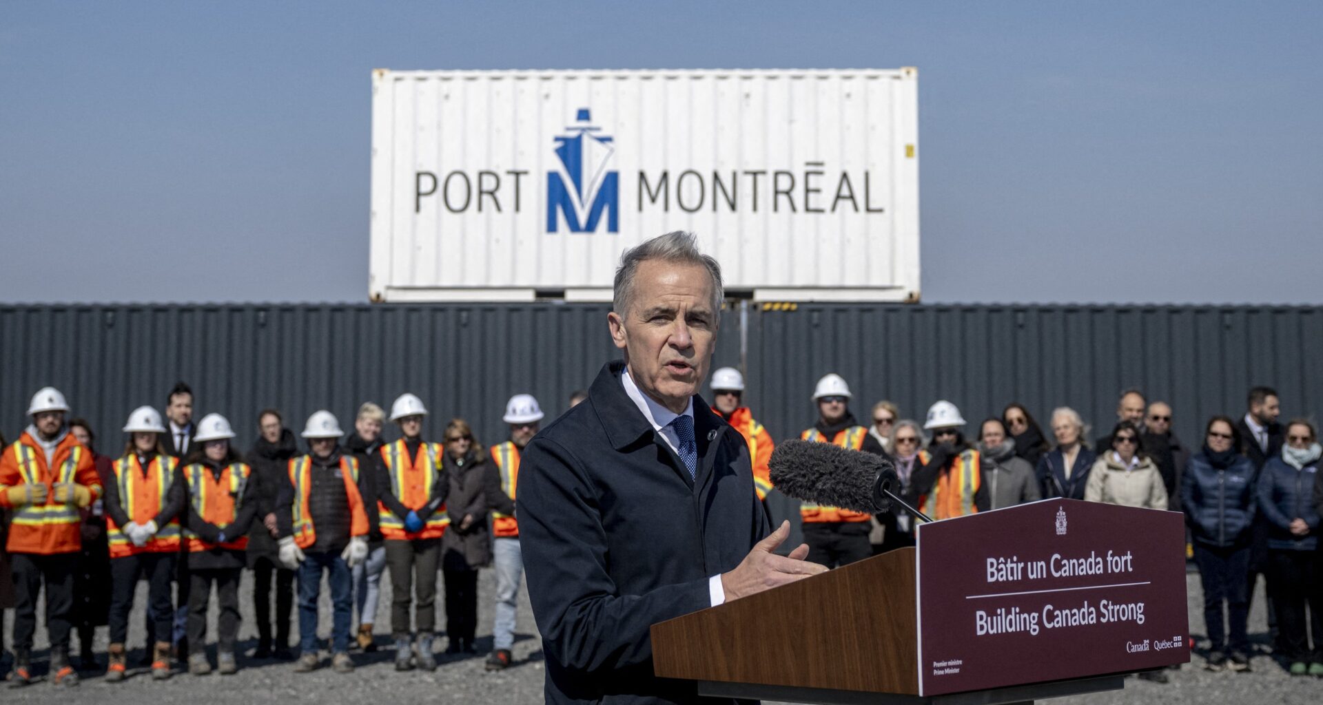 Prime Minister Mark Carney speaks at a podium with a bilingual sign on it reading Building Canada Strong. In the background is a Port of Montreal sign, a large port container and a row of people, many of them in construction uniforms.
