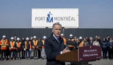 Prime Minister Mark Carney speaks at a podium with a bilingual sign on it reading Building Canada Strong. In the background is a Port of Montreal sign, a large port container and a row of people, many of them in construction uniforms.