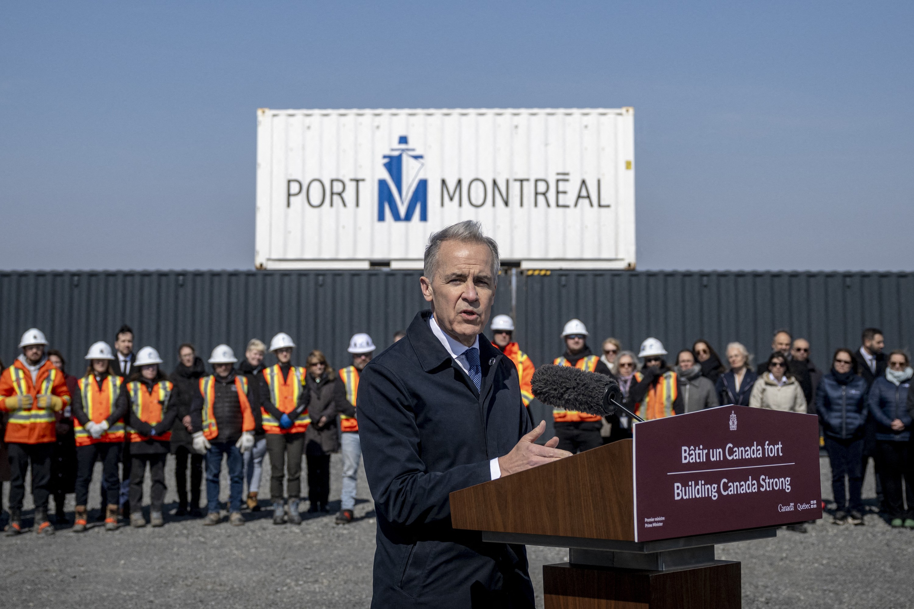 Prime Minister Mark Carney speaks at a podium with a bilingual sign on it reading Building Canada Strong. In the background is a Port of Montreal sign, a large port container and a row of people, many of them in construction uniforms.