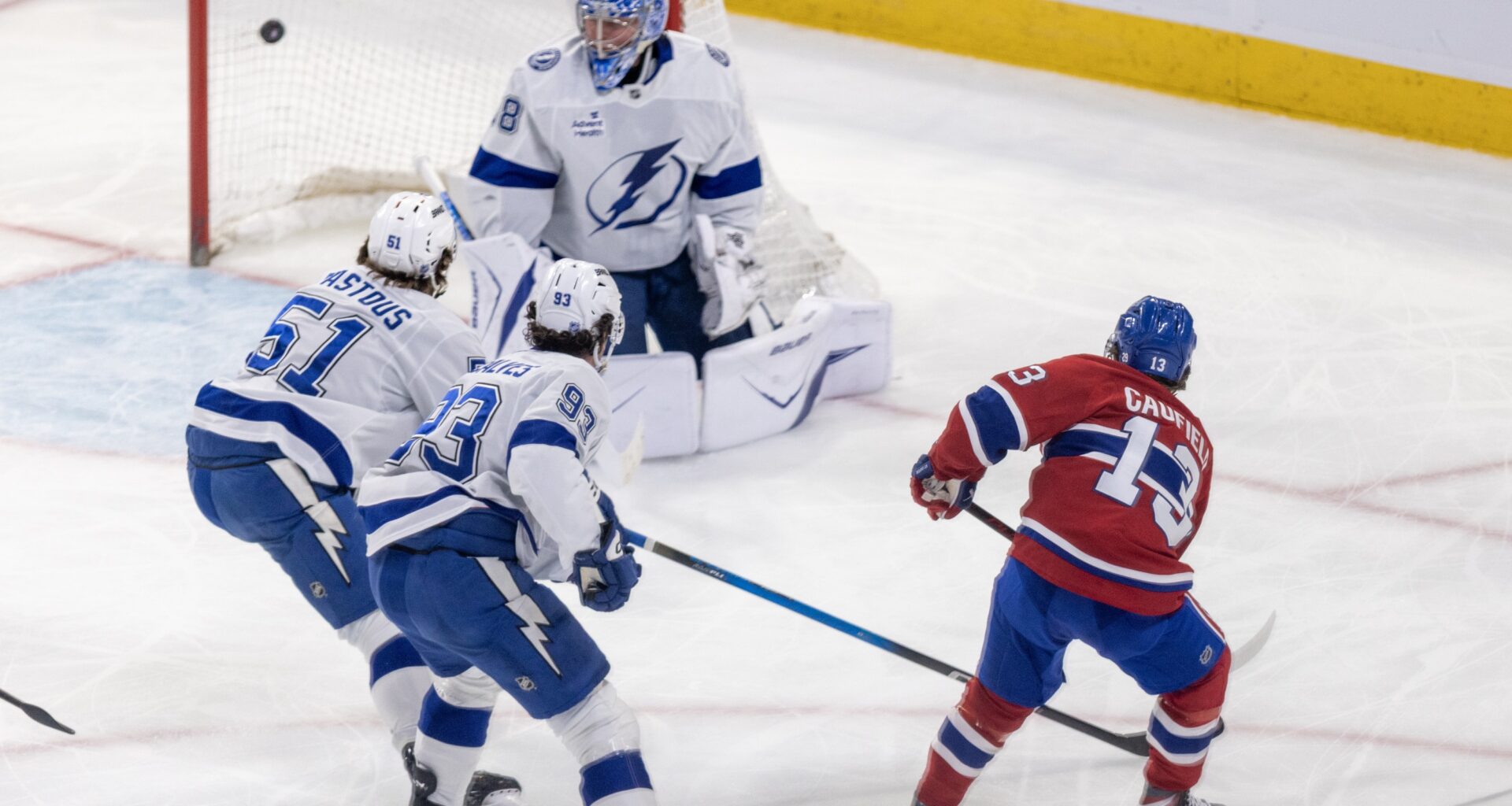 Hockey player shoots the puck past the goaltender with two other players defending.