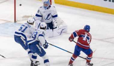 Hockey player shoots the puck past the goaltender with two other players defending.
