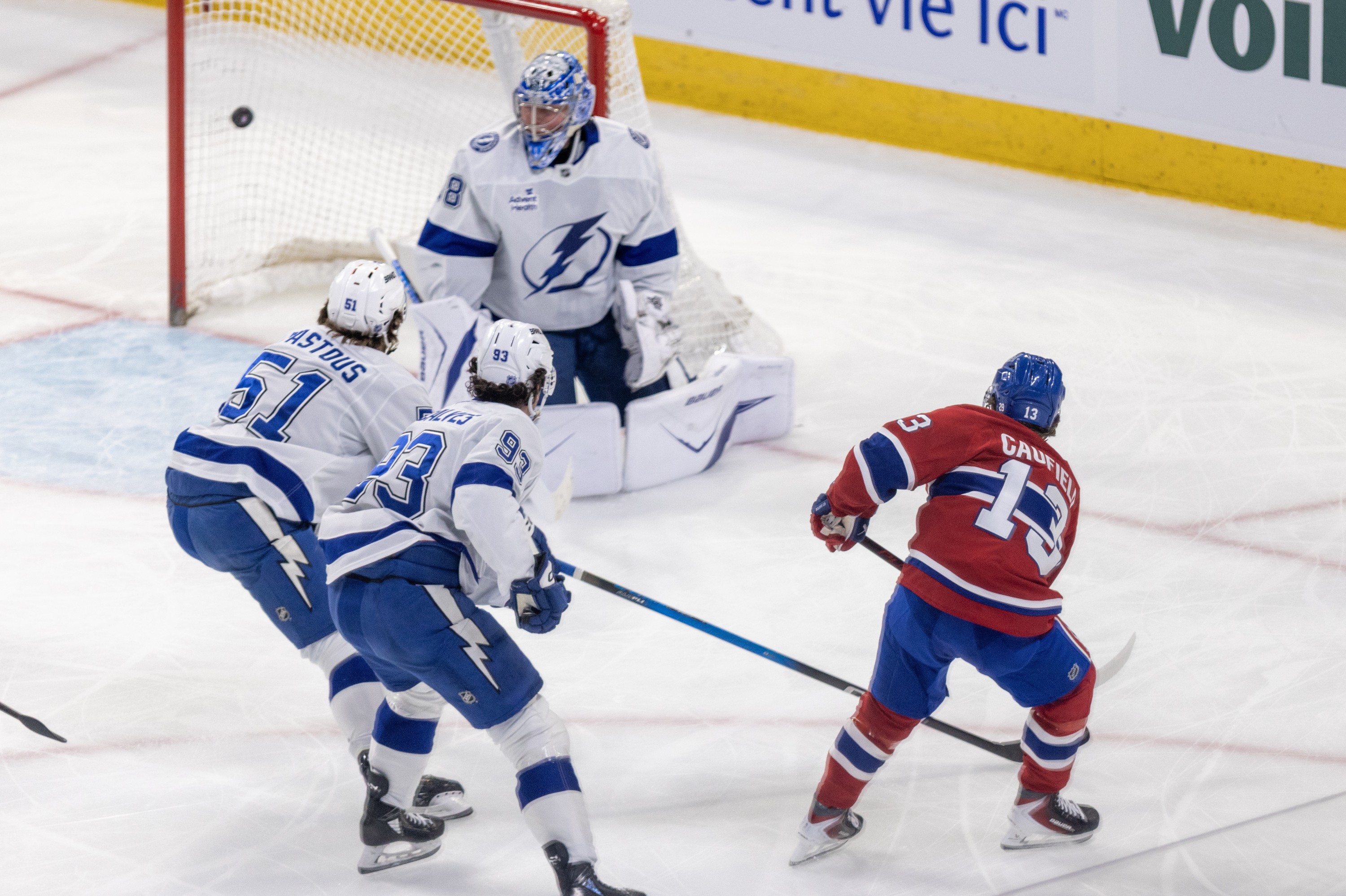 Hockey player shoots the puck past the goaltender with two other players defending.