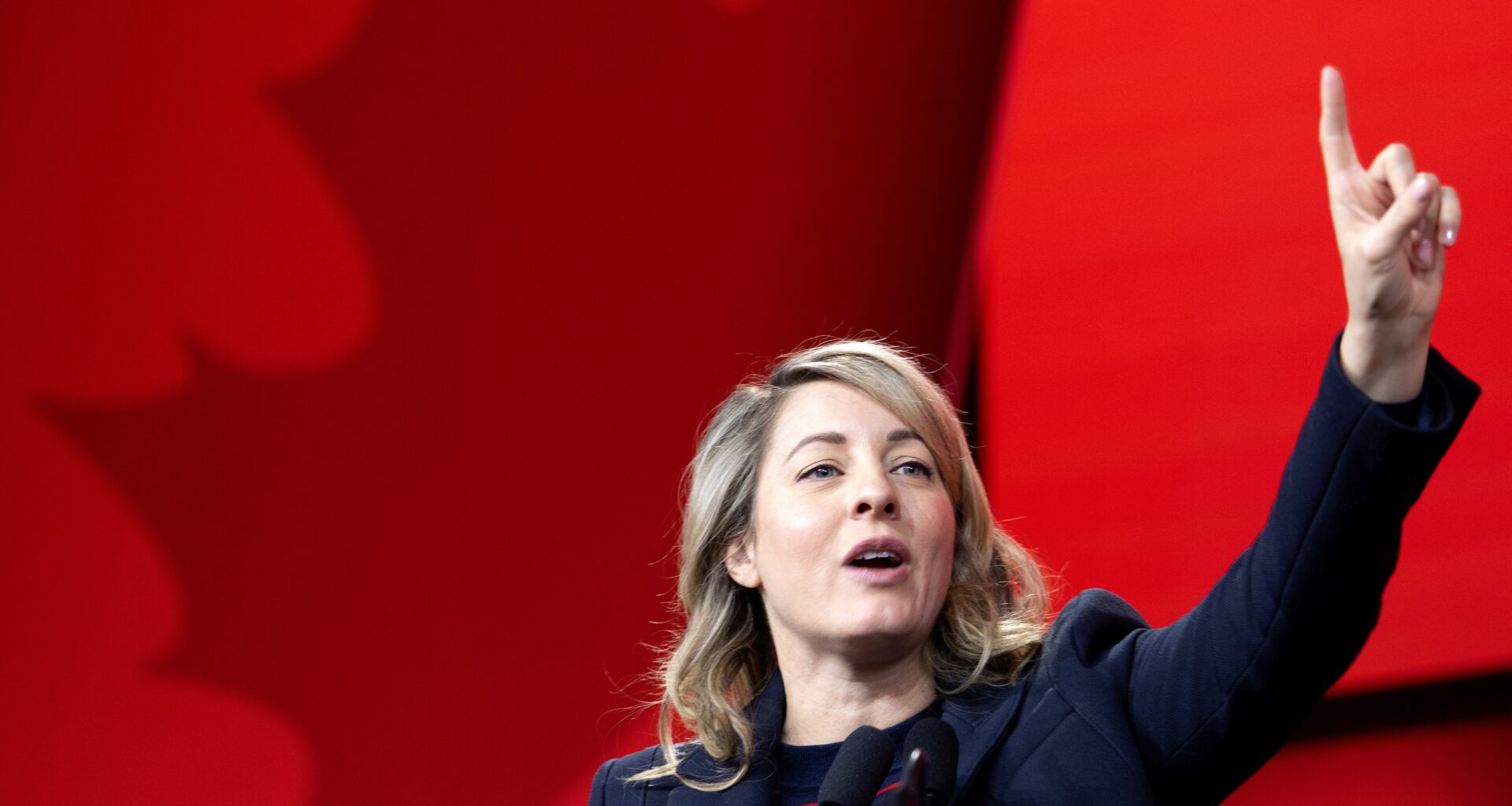 Female politician speaks at a podium with her arm raised and a finger held high with a red maple leaf background.