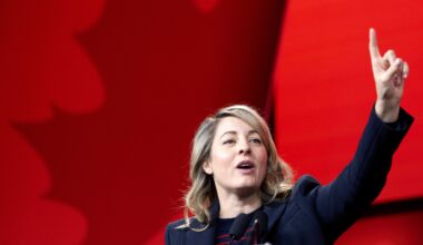 Female politician speaks at a podium with her arm raised and a finger held high with a red maple leaf background.