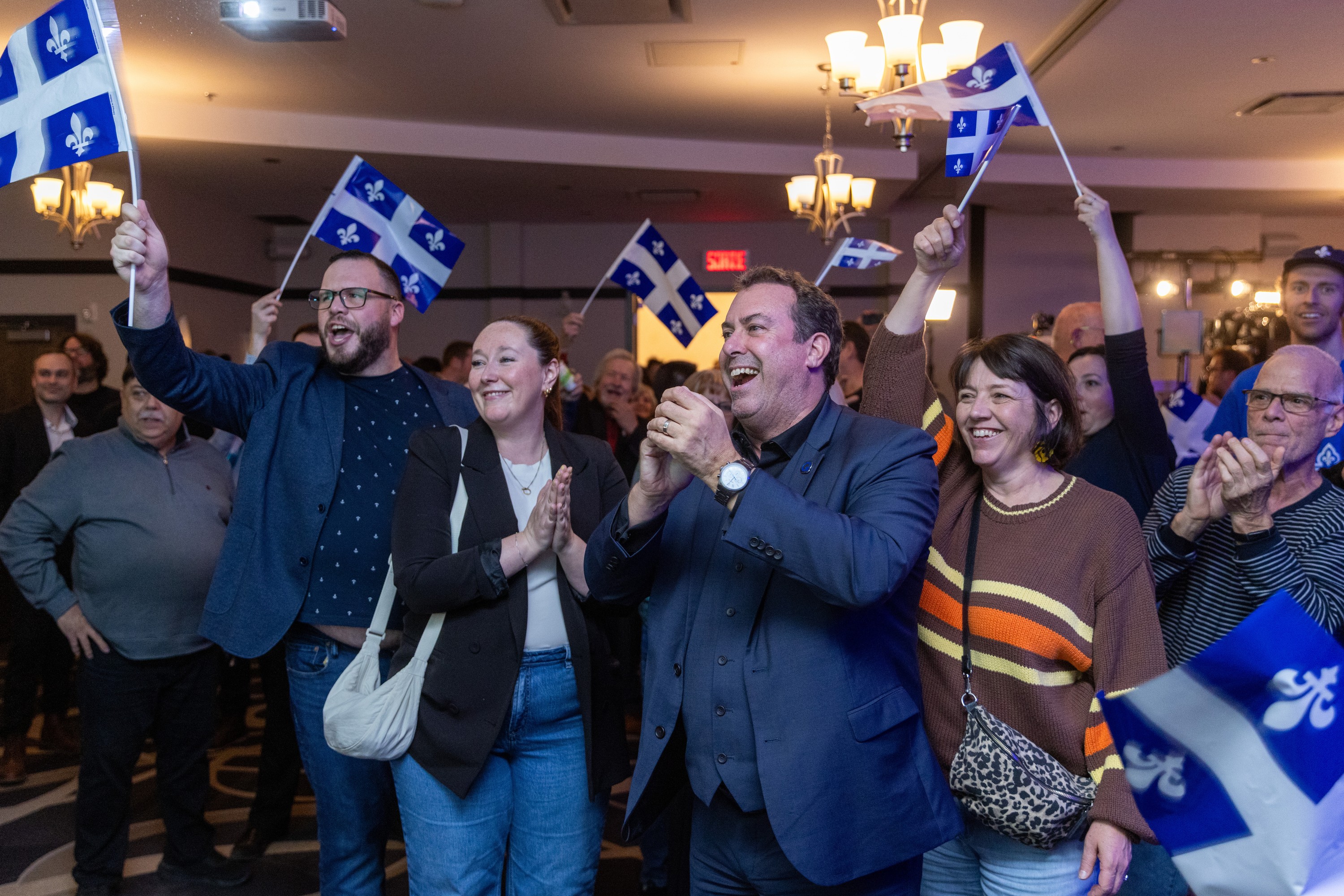People cheer holding Quebec flags in a large room.